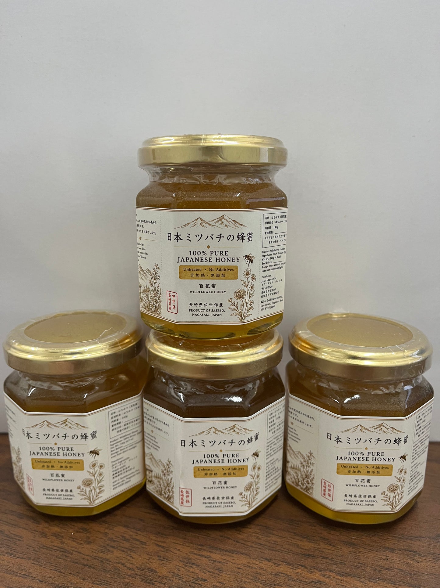 Four glass jars of Japanese honey with gold lids and cream labels, arranged on a wooden surface against a plain light background.