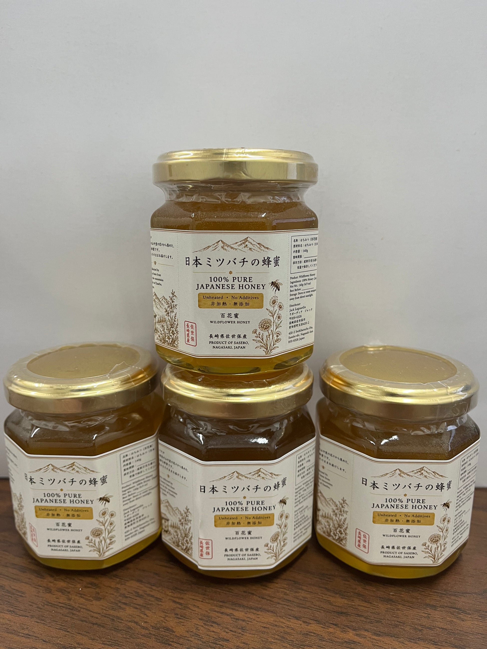 Four glass jars of Japanese honey with gold lids and cream labels, arranged on a wooden surface against a plain light background.