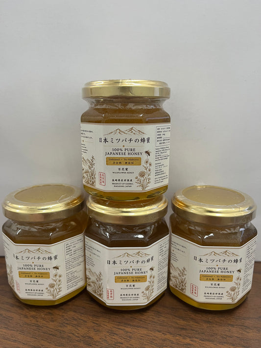 Four glass jars of Japanese honey with gold lids and cream labels, arranged on a wooden surface against a plain light background.